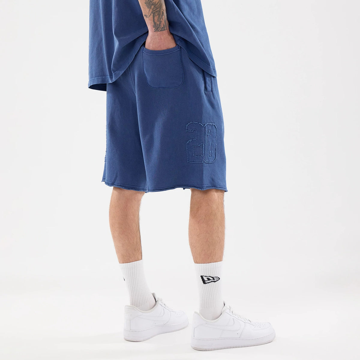 New Era Street Archived Blue Oversized Shorts, side view showing the blue fabric with a debossed number "28" on the left leg, a wide pocket at the upper side, worn with white socks featuring a New Era logo above white trainers.