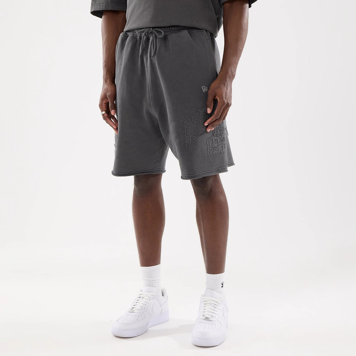 New Era Street Archived Black Oversized Shorts, front view showing charcoal grey shorts with a raised New Era logo on the left thigh, an elasticated waistband with a drawstring and side pockets, worn with white trainers.