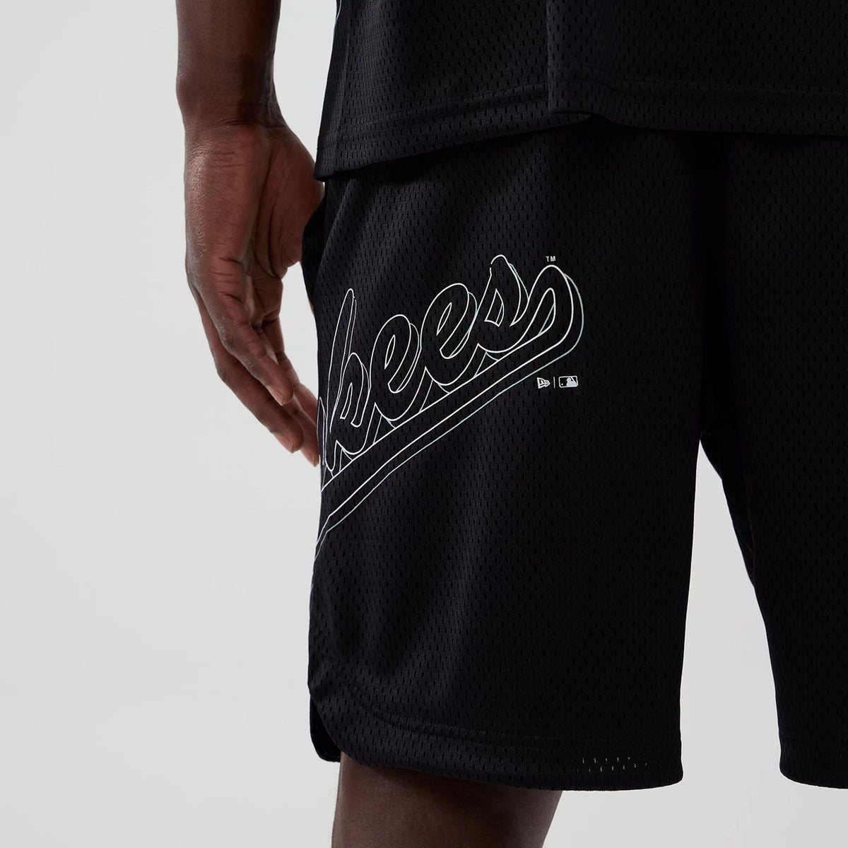 New York Yankees MLB Mix League Black Oversized Mesh Shorts, side view showing the black mesh fabric with a raised white Yankees script logo near the hem and a hand resting lightly on the seams.