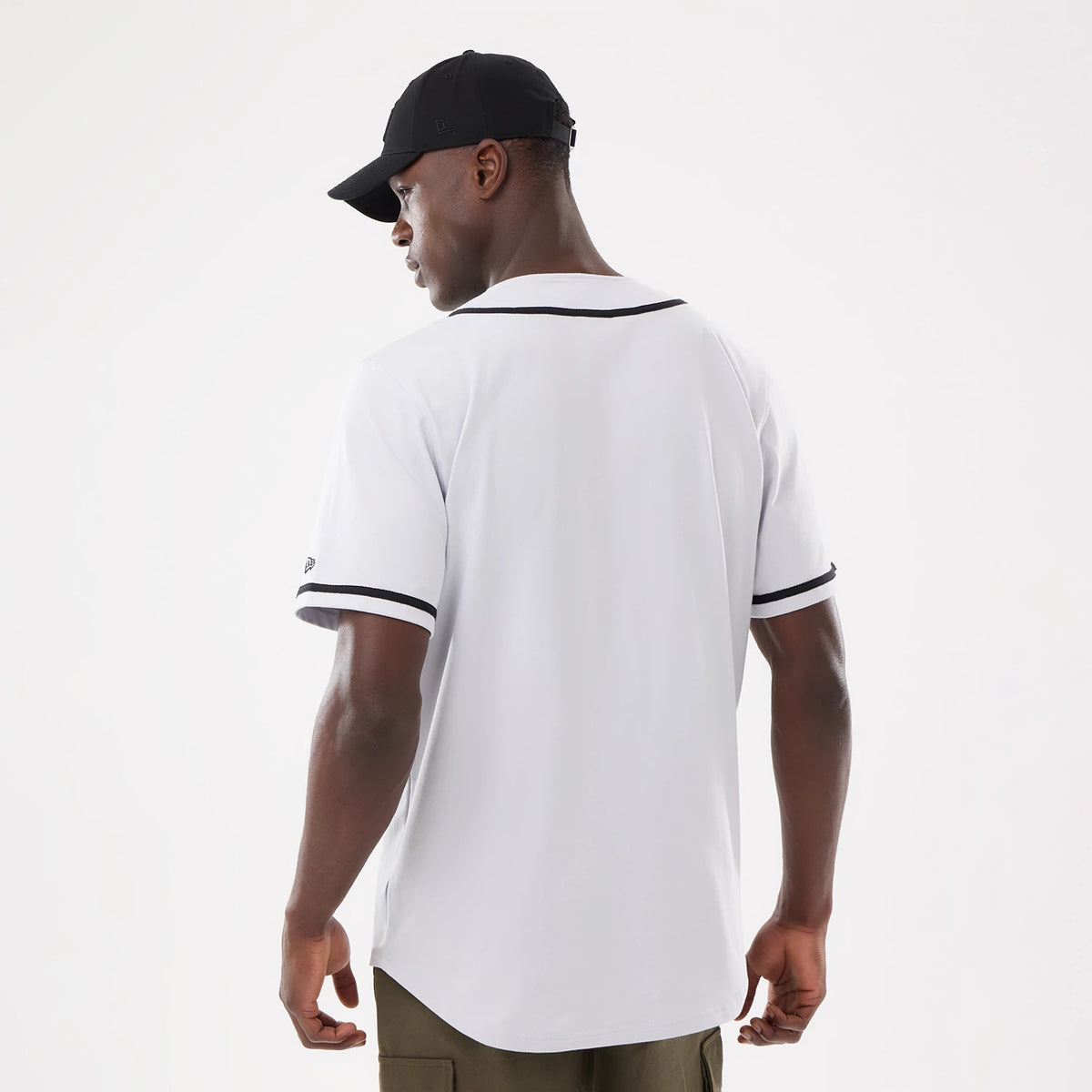 New Era Basic White Jersey, back view on a model wearing a black cap, highlighting black piping around the collar and sleeves, along with a relaxed fit that drapes over olive trousers.