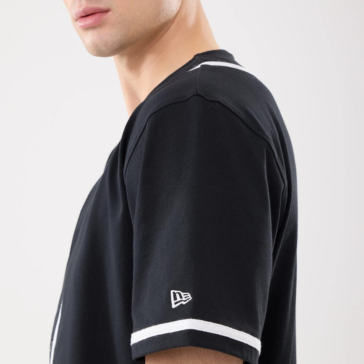 New Era Basic Black Jersey, side view on a model showing the white New Era flag logo embroidered on the left sleeve and contrasting white trim along the sleeve end, highlighting the clean lines of the black fabric.