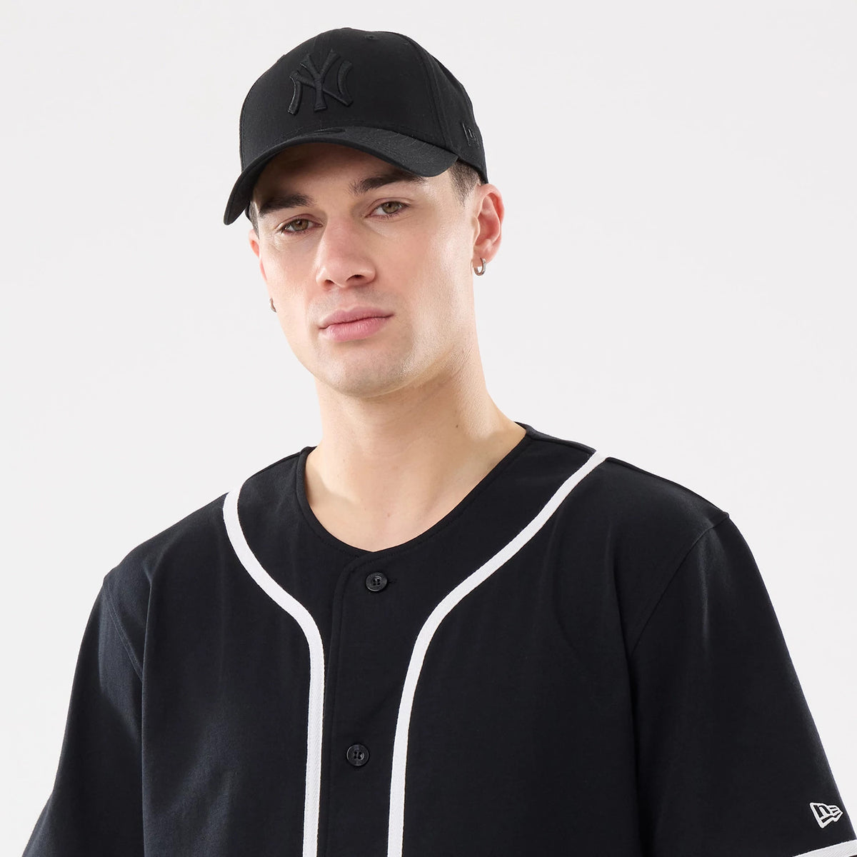 New Era Basic Black Jersey, on-model front view with a black cap featuring a raised black NY logo on the front panel, worn with a matching black jersey outlined with white piping and a New Era flag logo on the sleeve.