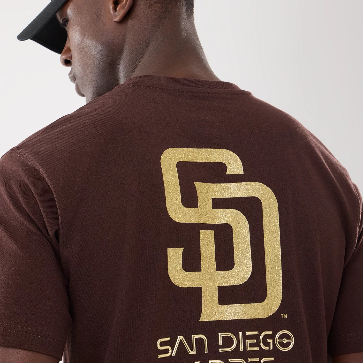 San Diego Padres Hajime Sorayama X MLB Dark Brown T-Shirt, rear view showing a large embossed gold SD logo centred on the back, worn by a model in a black cap, with the embossed text "SAN DIEGO PADRES" below the logo.