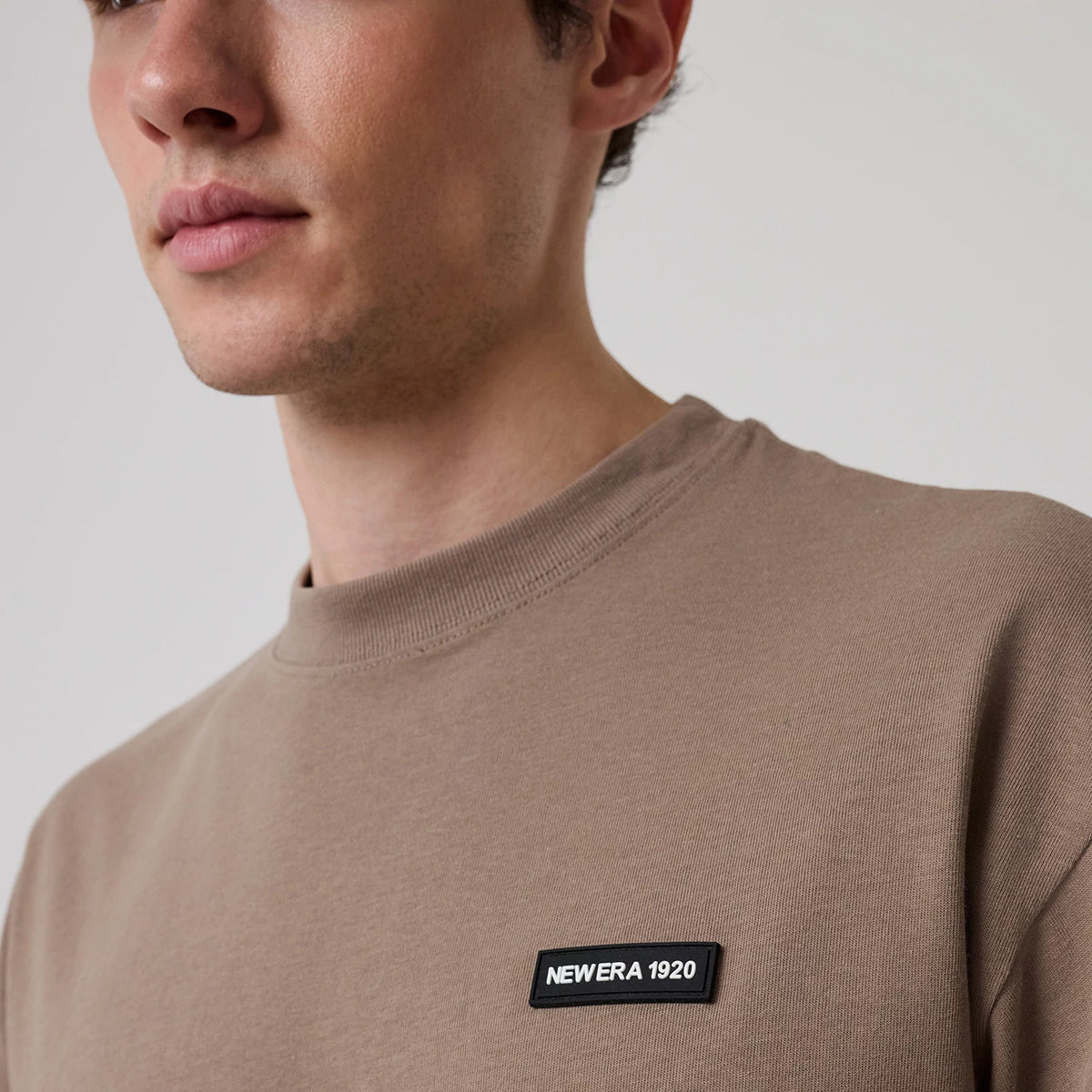 New Era Distressed Brown T-Shirt, on-model view showing the light brown fabric with a raised black and white New Era 1920 logo patch on the left chest, featuring a close-up of the ribbed crew neckline.