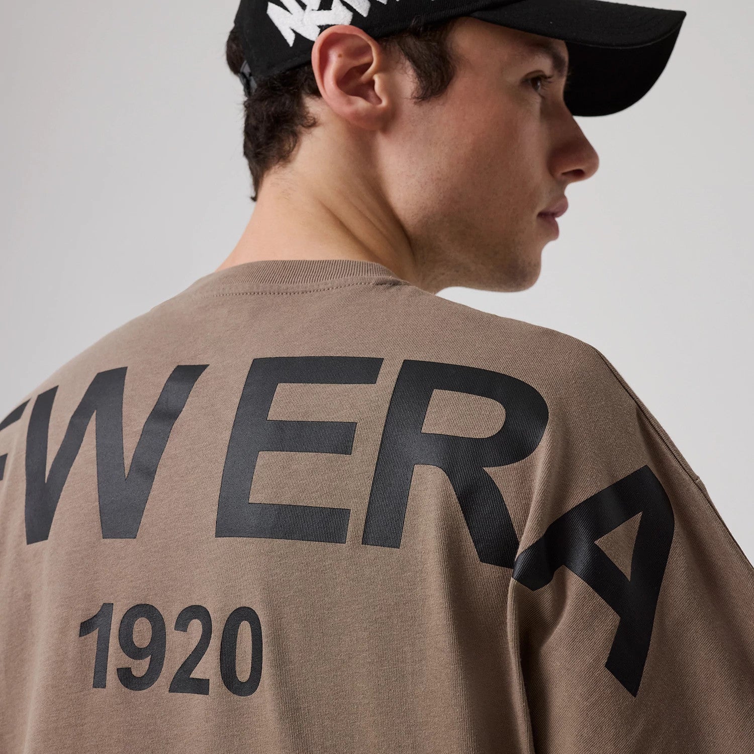 New Era Distressed Brown T-Shirt, on-model view showing the large black printed letters 'NEW ERA' and '1920' across the back, with a black cap featuring a white raised logo worn by the model.