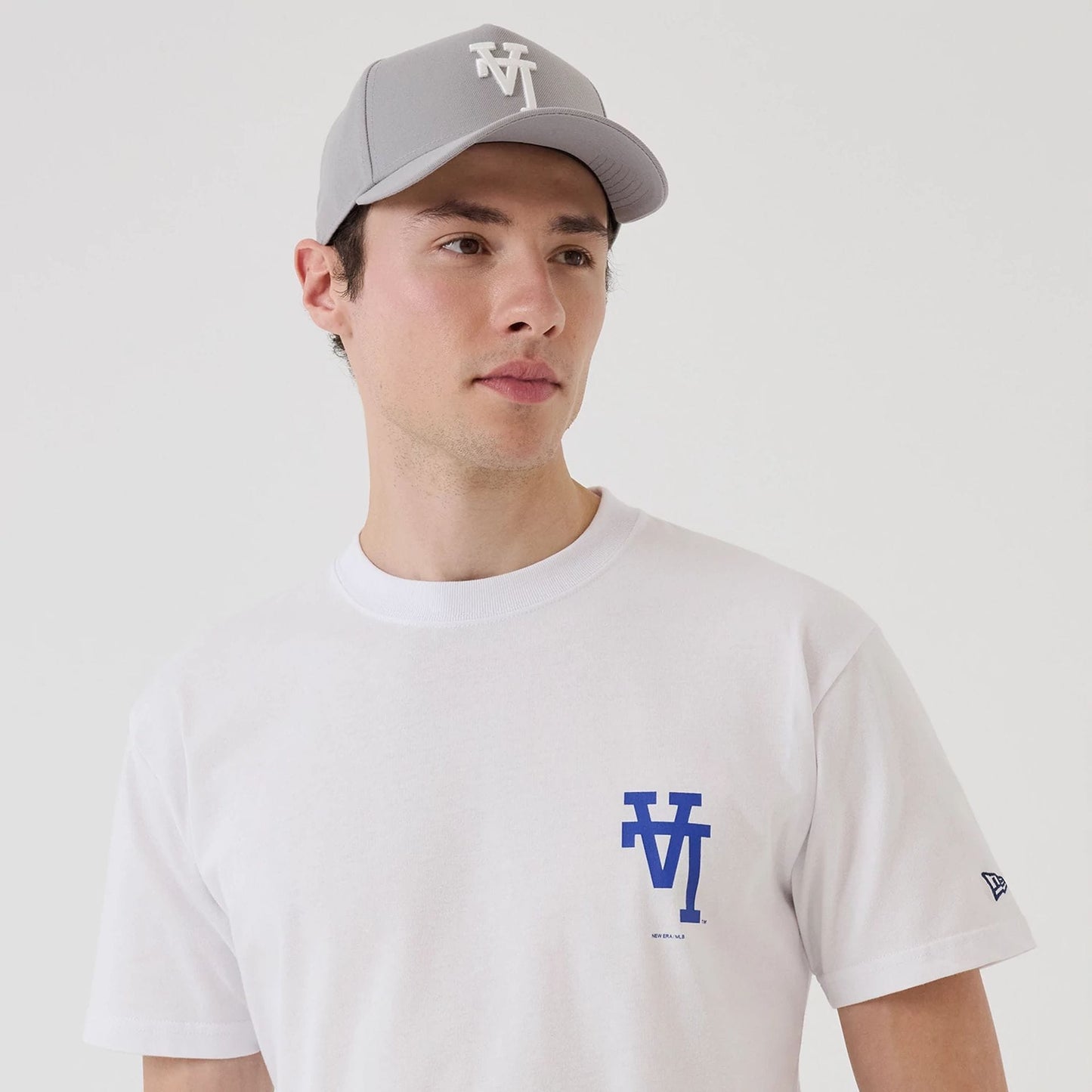 LA Dodgers MLB Upside Down White T-Shirt, on-model view showing the blue logo printed on the left chest of a plain white T-shirt and a grey cap with a raised logo on the front worn by the model.