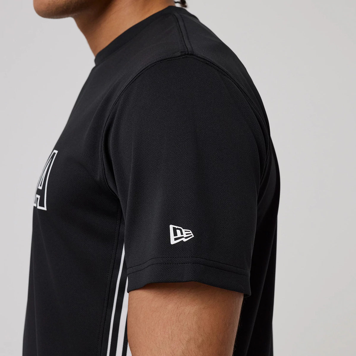 New Era Mesh Black T-Shirt, side view showing a close-up of the short sleeve with a raised New Era logo on the cuff, contrasted against the black fabric, and white design elements running down the side.
