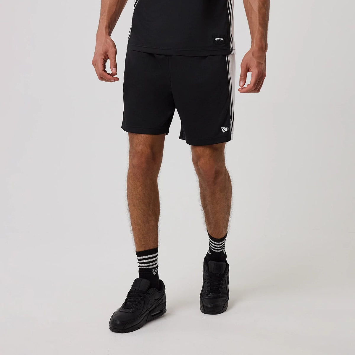 New Era Mesh Black Shorts, on-model view showing the black shorts with a white striped design on the sides, an embroidered New Era logo near the hem on the left leg, worn with matching black trainers and striped socks.