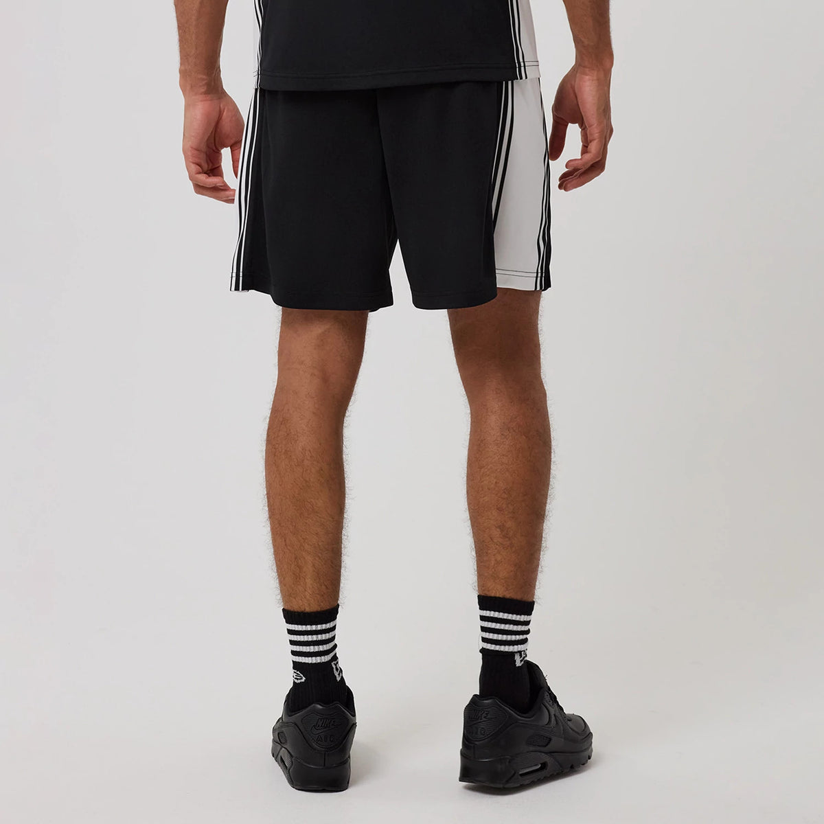 New Era Mesh Black Shorts, rear view showing the black shorts with white side panels and vertical stripes, worn on-model with black trainers and striped socks, highlighting the contrast between the black and white elements.