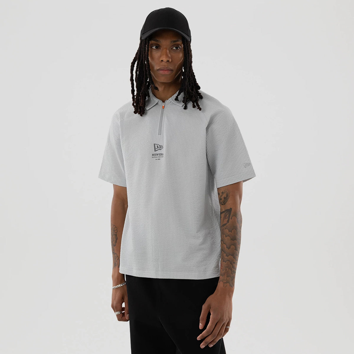 New Era Seersucker Grey Polo T-Shirt, on-model view featuring a light grey textured polo with a half zip and a raised New Era logo centred on the chest, short sleeves, and a casual fit worn by a person in black trousers.