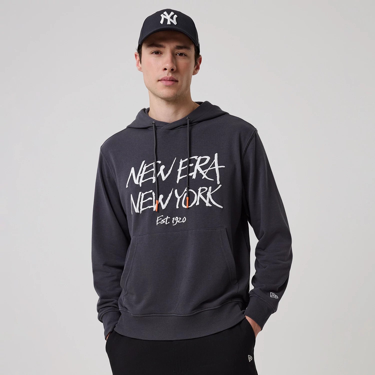 New Era New York Dark Grey Oversized Pullover Hoodie, on-model view showing the front graphic with "New Era New York Est 1920" in white, a debossed New Era logo on the left sleeve cuff, and a drawstring hood.