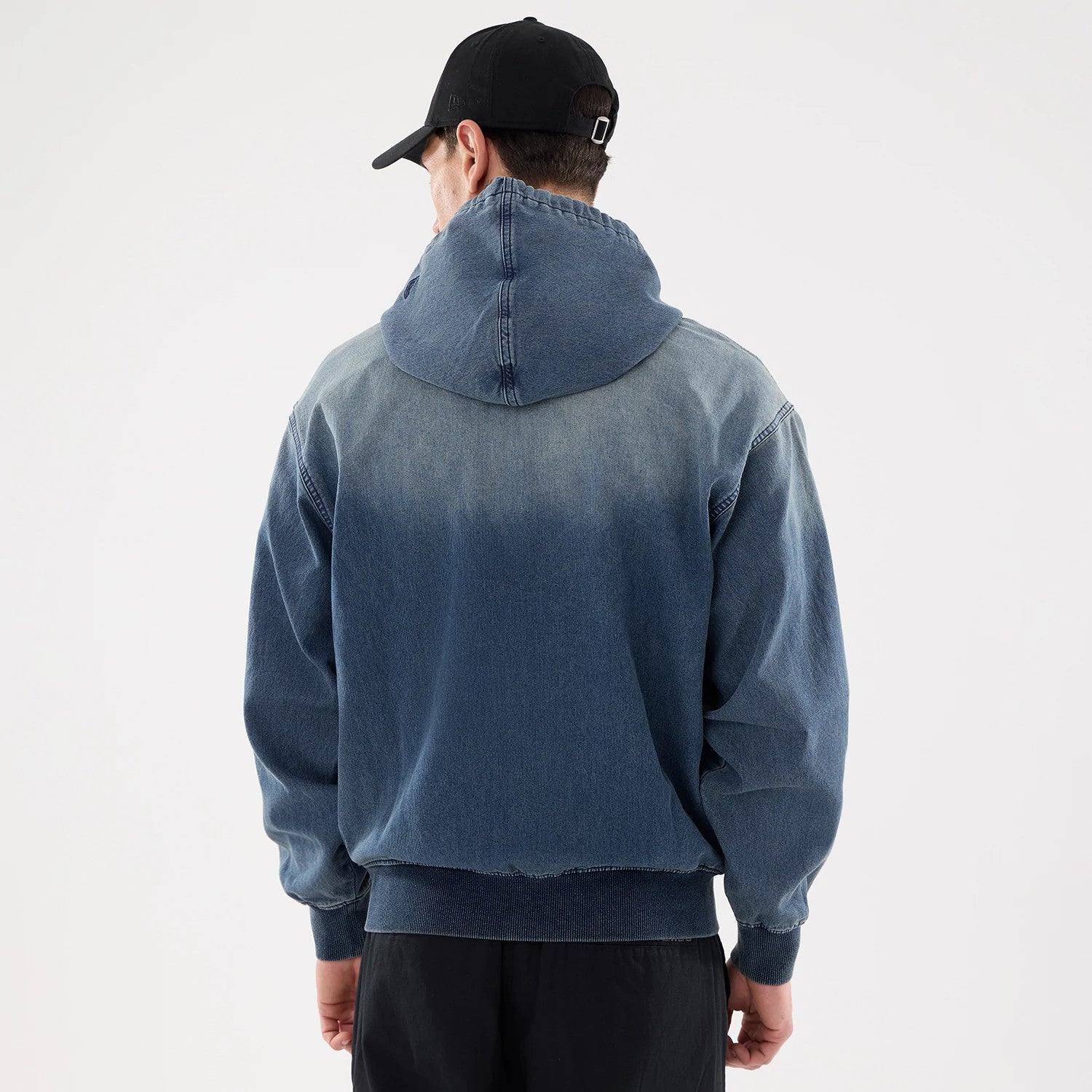 New Era Washed Blue Full Zip Hoodie, rear view showing the faded blue back with a hood, ribbed cuffs and hem, worn by a model in black trousers, and a black cap with a debossed logo on the back.