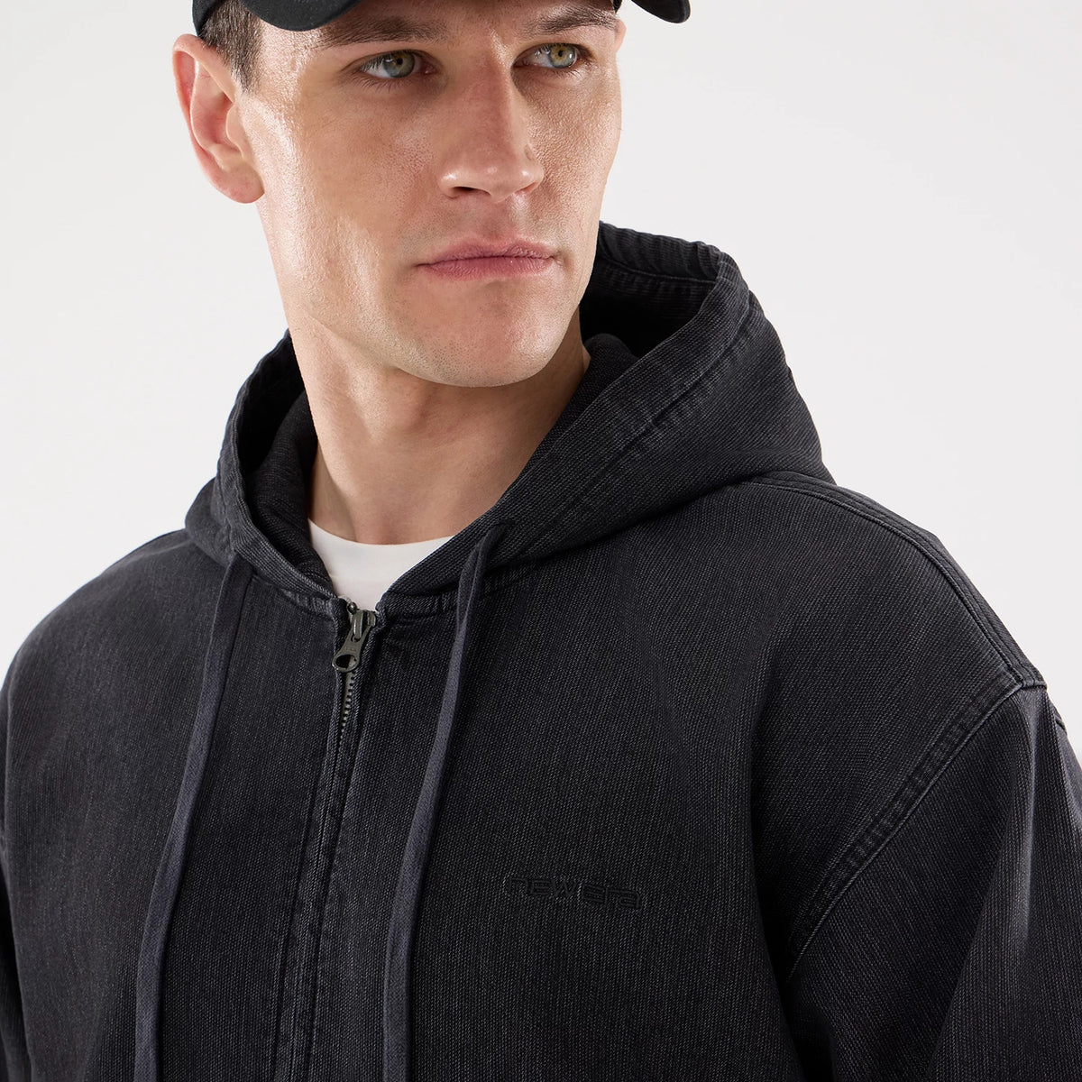 New Era Washed Black Full Zip Hoodie, three-quarter view showing the debossed logo on the left chest, a full-length zip running down the front, and an attached hood with drawstrings, worn by a man in a black cap.