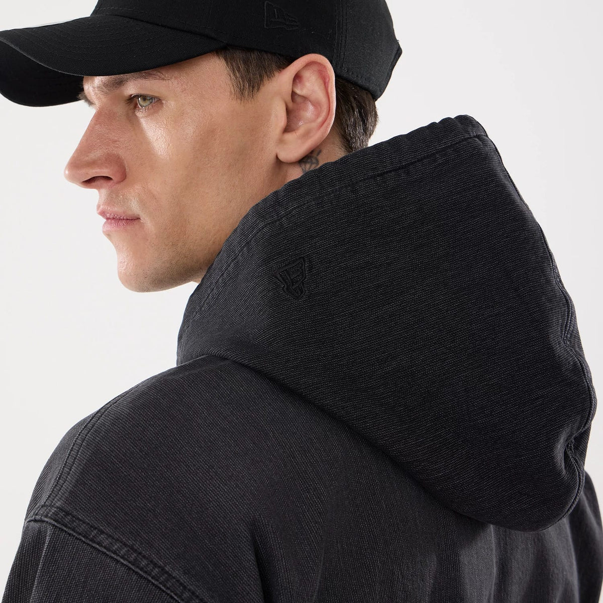 New Era Washed Black Full Zip Hoodie, side view showing the washed black hood with a debossed New Era logo on the side, worn over a black cap with a raised New Era logo on the side.