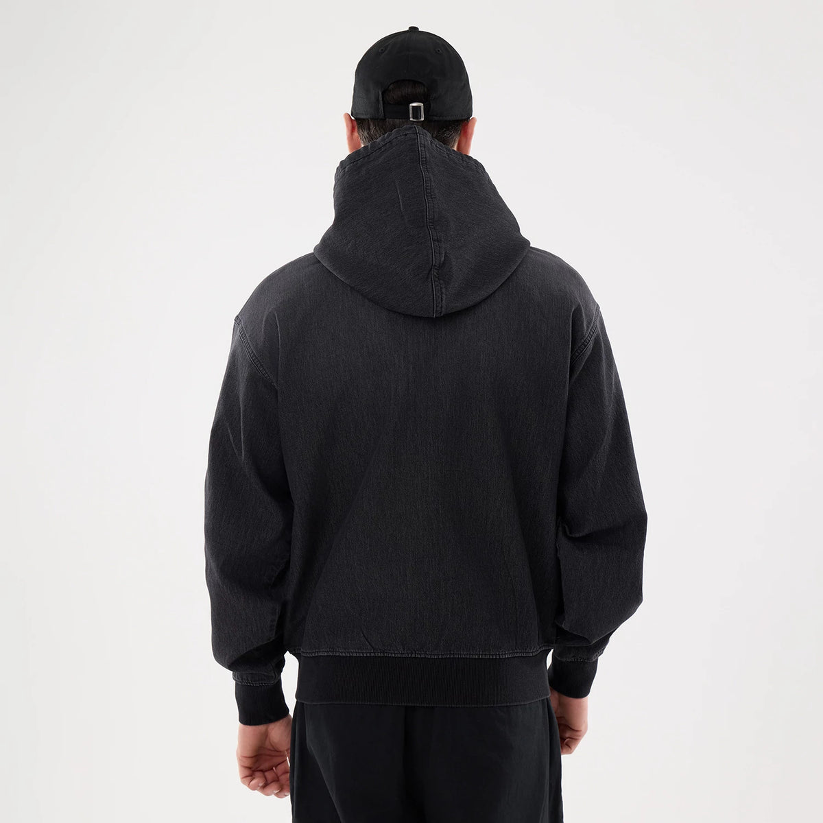 New Era Washed Black Full Zip Hoodie, rear view showing the hooded design with relaxed shoulders and ribbed cuffs, worn by a model also wearing a black cap with a debossed logo on the back.