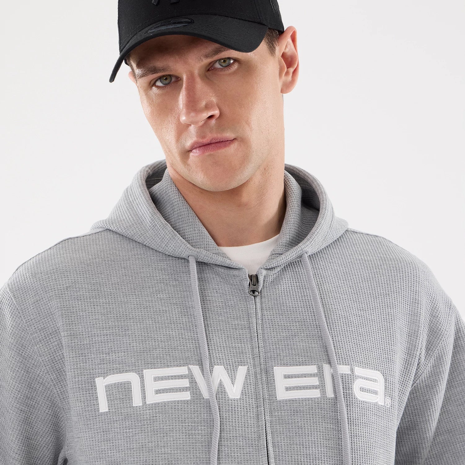 New Era Waffle Grey Full Zip Hoodie, on-model view showing the light grey textured fabric with a full-length zip, the hood resting on the shoulders, and a large white New Era logo printed across the chest.