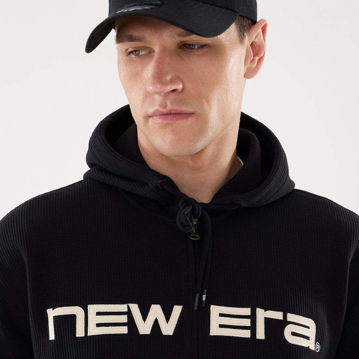 New Era Waffle Black Full Zip Hoodie, on-model view showing the textured black fabric with a large white embroidered 'New Era' logo across the chest and a drawstring hood, paired with a black cap featuring a raised logo on the front.