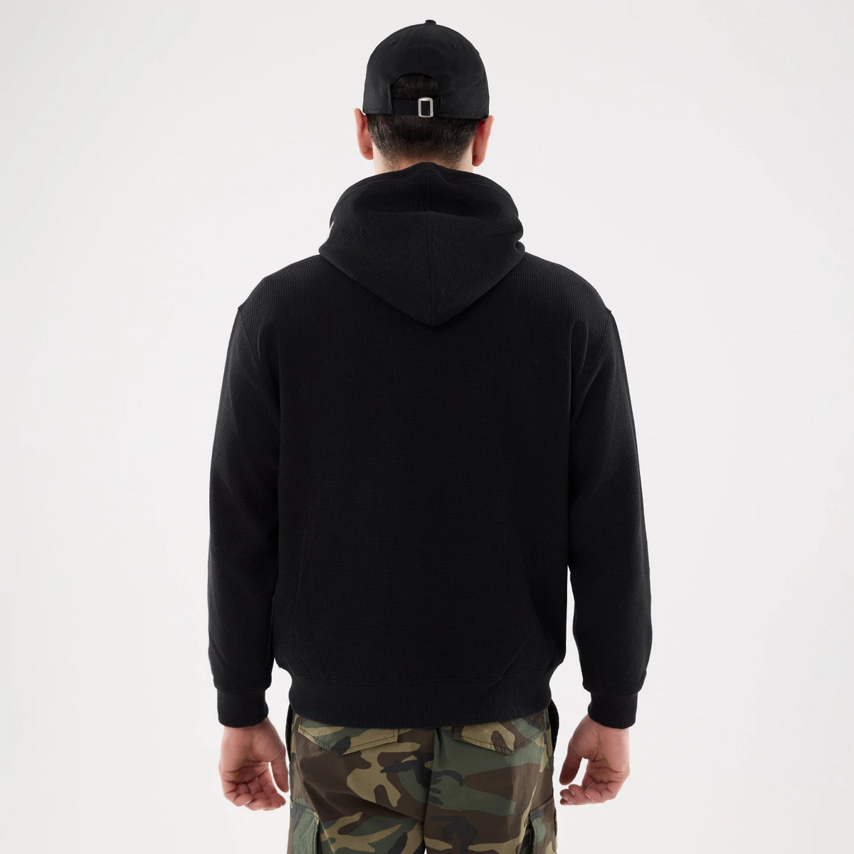 New Era Waffle Black Full Zip Hoodie, rear view showing the textured black fabric and hood at the back, worn by a model in camouflage trousers and a black cap with an adjustable strap.