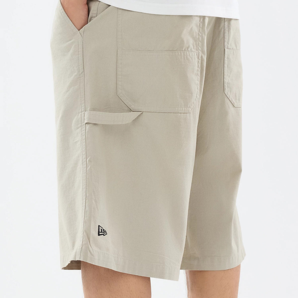 New Era Canvas Beige Bermuda Shorts, side view showing the straight leg cut and large patch pockets on the side, a small flap detail near the pocket, and an embroidered New Era logo near the hem.