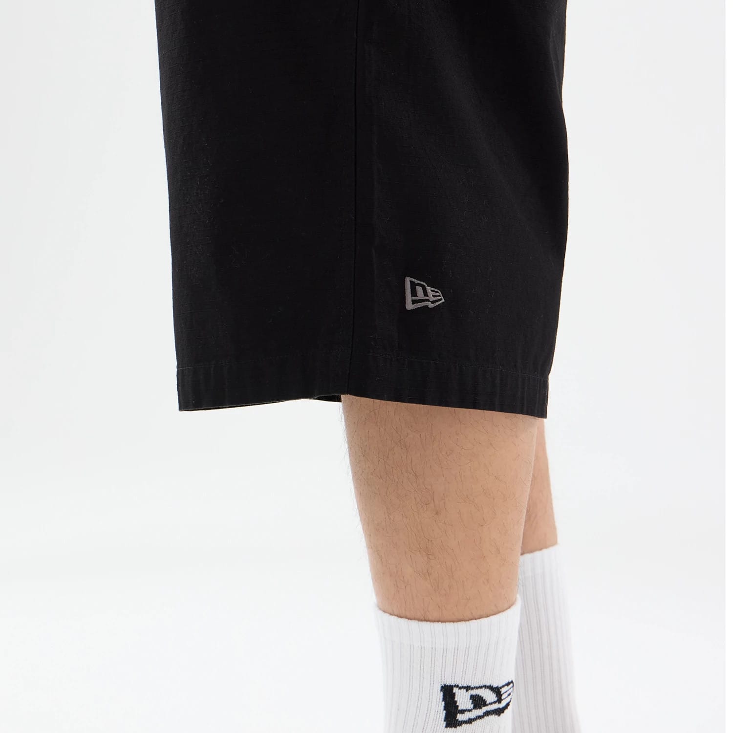 New Era Canvas Black Bermuda Shorts, on-model view showing the lower section of the black shorts, a debossed New Era logo near the hem and white socks with a New Era logo pattern on the model's legs.