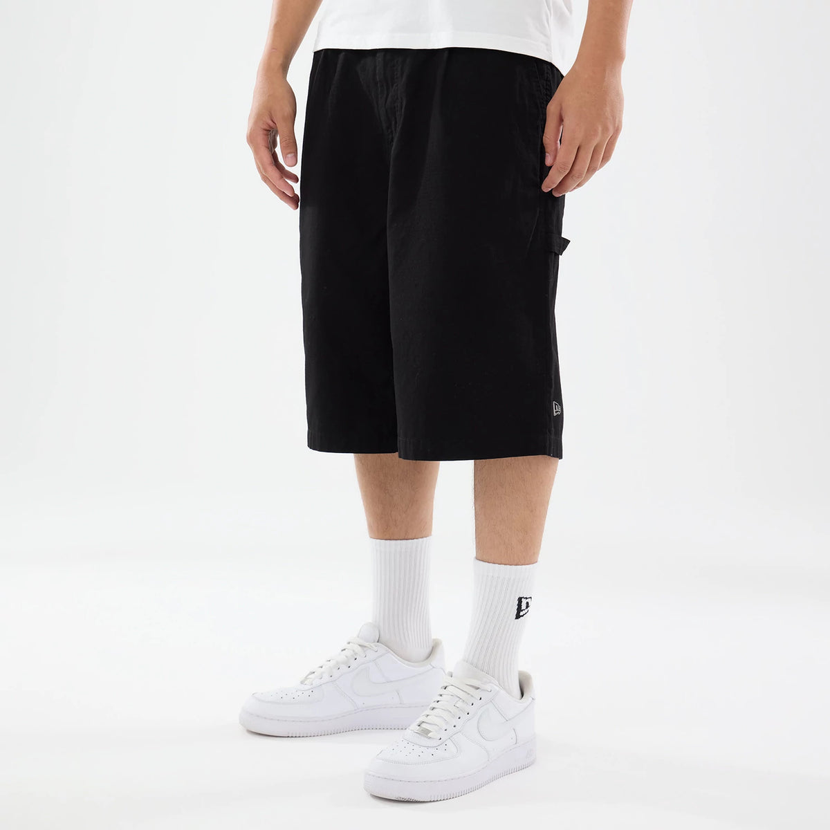 New Era Canvas Black Bermuda Shorts, on-model view featuring knee-length black shorts with a relaxed fit and a raised New Era logo on the lower left leg, paired with white crew socks and white trainers.