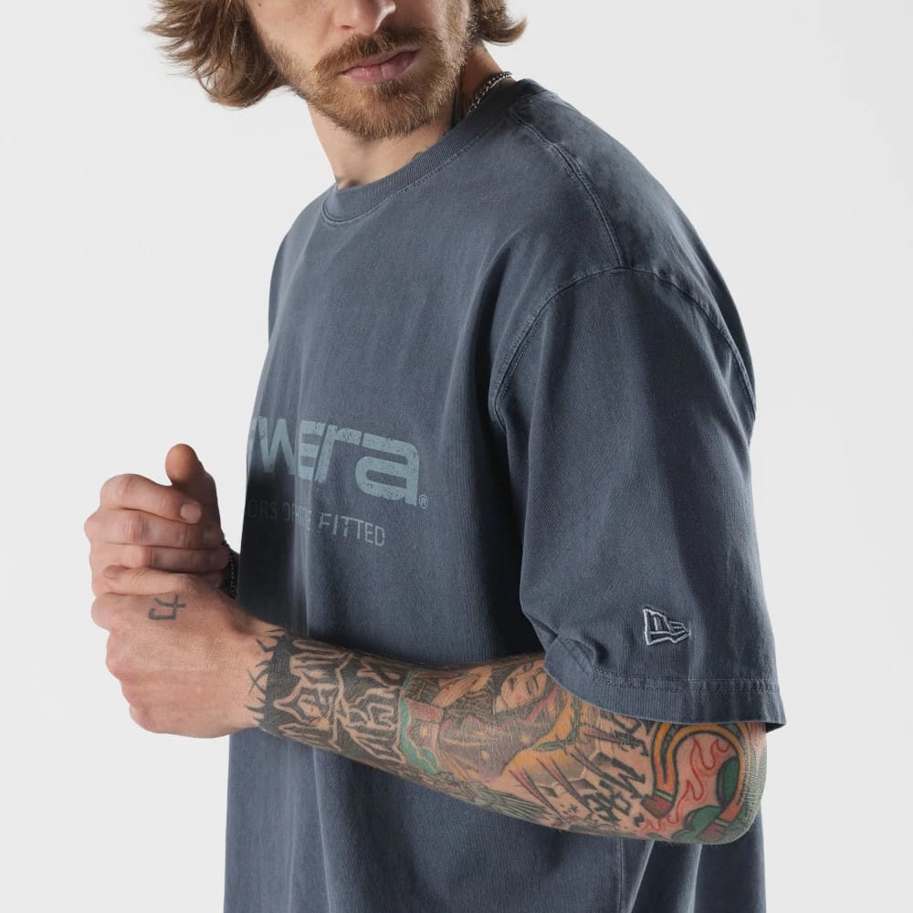 New Era Originators Navy T-Shirt, on-model view showing a navy T-shirt with a large embossed New Era logo across the chest, short sleeves with a smaller embossed logo near the hem on one sleeve, worn by a person with tattooed arms.