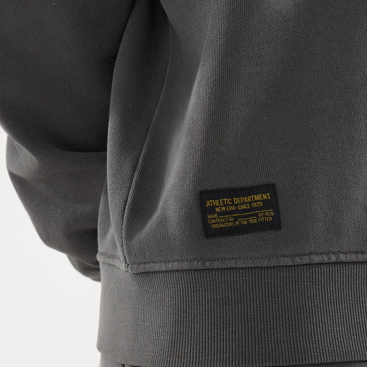 New Era Originators Black Crew Neck Sweater, close-up showing the yellow text patch on the right side of the ribbed hem, featuring "ATHLETIC DEPARTMENT" and "ORIGINATORS OF THE TRUE FITTED" embroidery.
