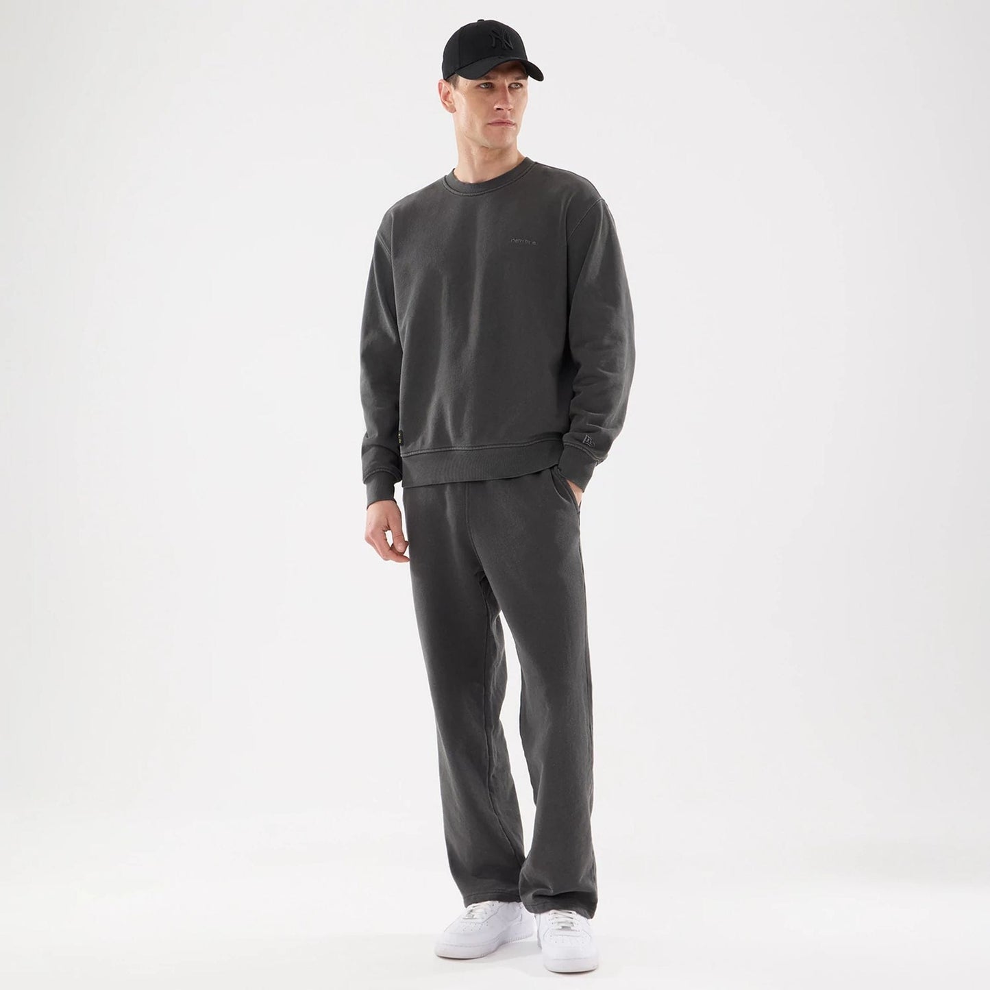 New Era Originators Black Crew Neck Sweater, on-model with matching dark trousers and a black cap, showing the embroidered Originators logo on the chest and the New Era flag logo on the left sleeve.