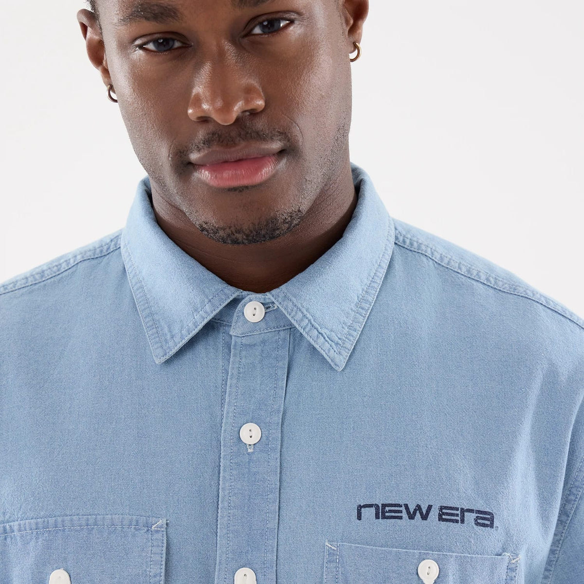 New Era Originators Chambray Bright Blue Short Sleeve Shirt, front view on a model showing the button-down collar, embroidered New Era logo above the left chest pocket, and bright blue chambray fabric.