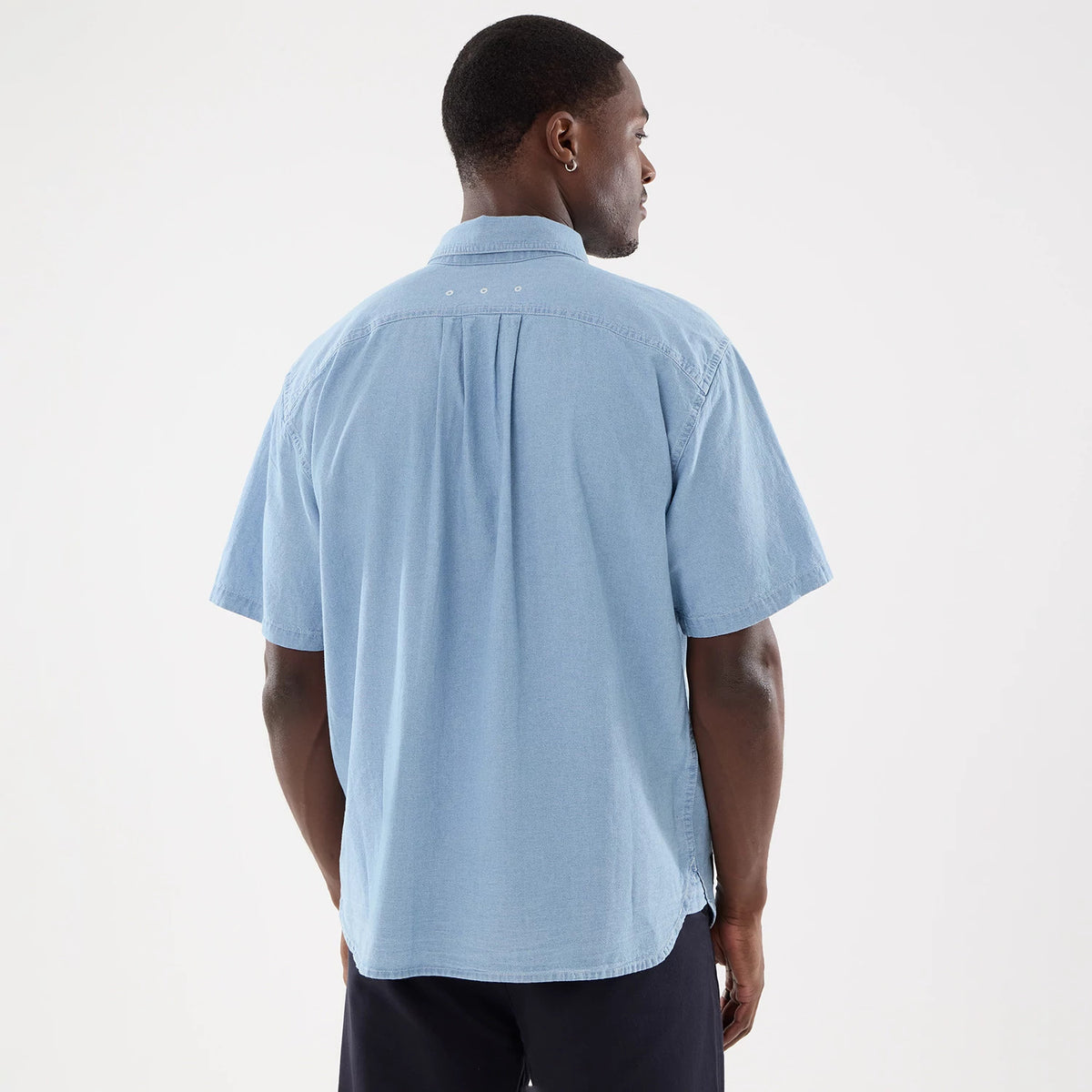 New Era Originators Chambray Bright Blue Short Sleeve Shirt, back view on a model wearing dark trousers, showing pleat and tab details on the upper back and a relaxed fit with a straight hemline.