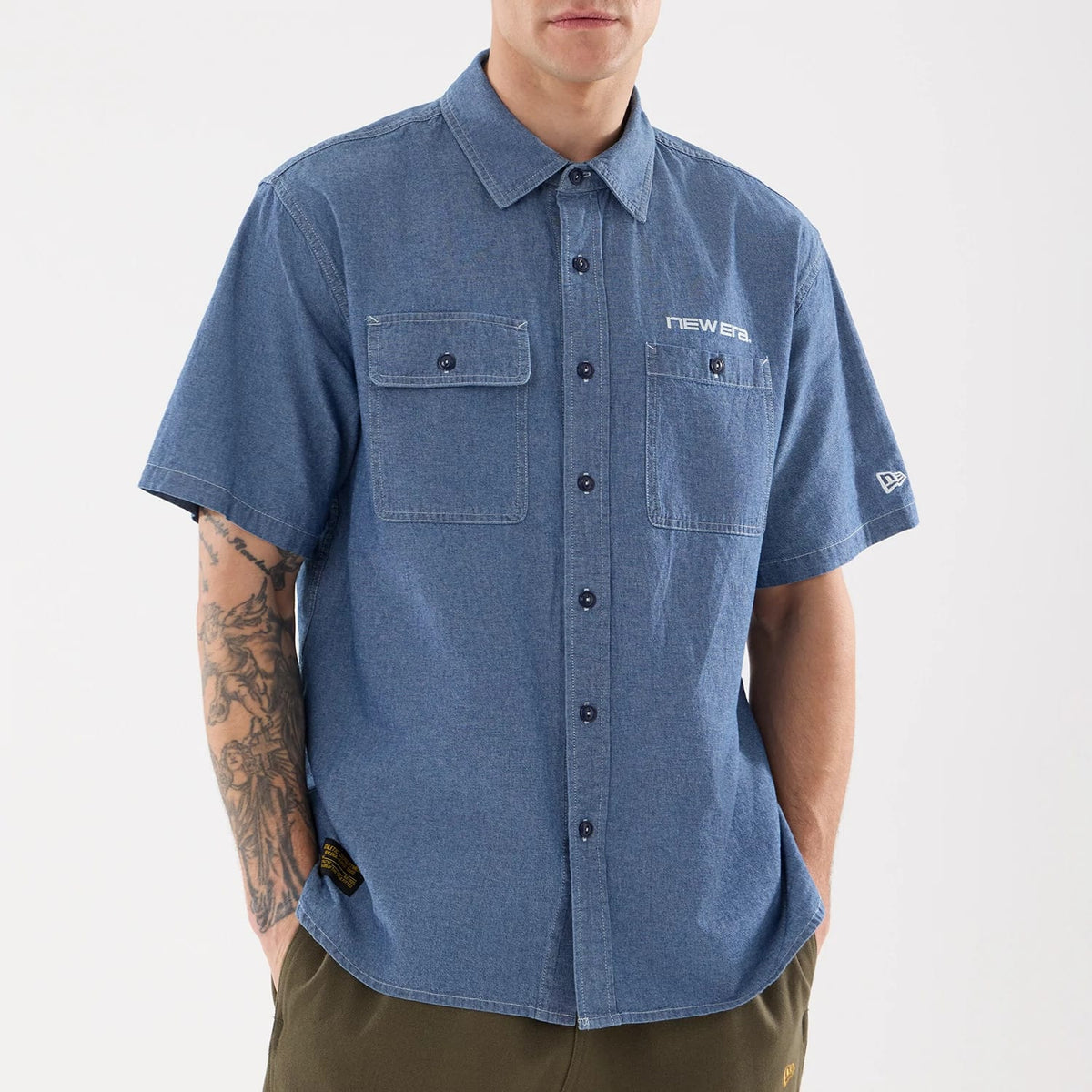New Era Originators Chambray Dark Blue Short Sleeve Shirt, front view on a model showcasing button-down front, chest pockets with button closure, and embroidered New Era logo on the left chest and sleeve, styled with olive trousers.