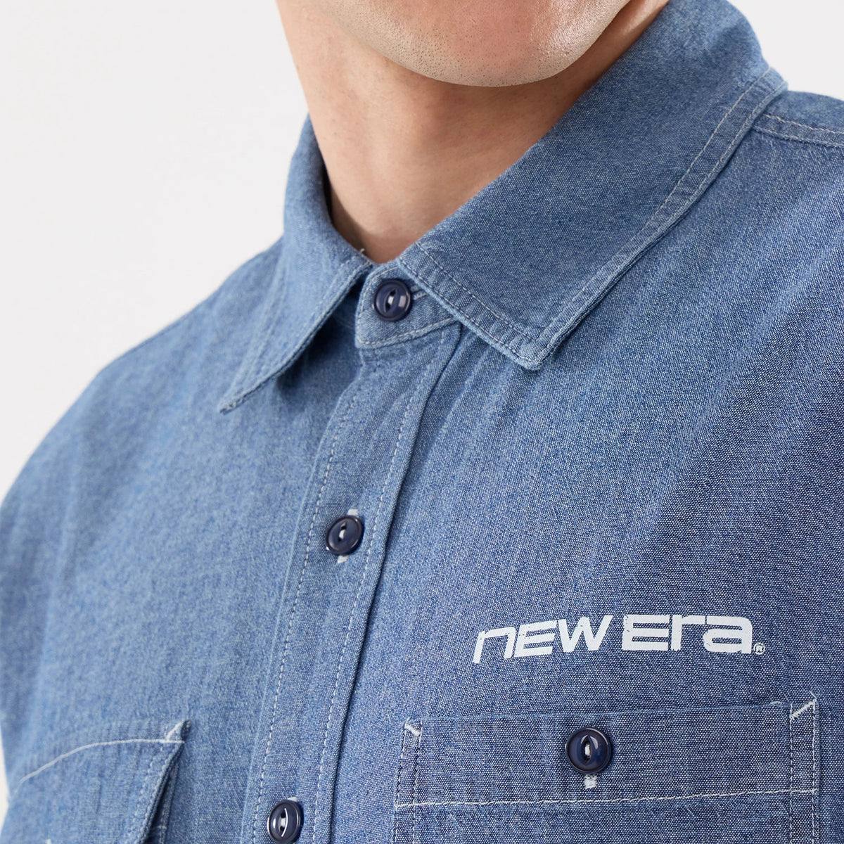 New Era Originators Chambray Dark Blue Short Sleeve Shirt, close-up showing white New Era lettering above the left chest pocket, with dark blue buttons and a classic collar design on a model.