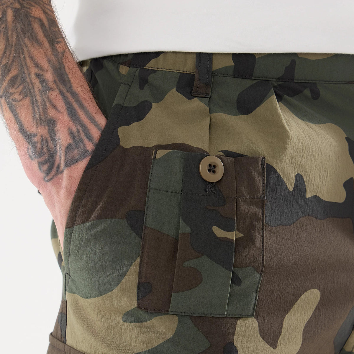 New Era Originators Woodland Camo Cargo Trousers, close-up view showing the detailed woodland camouflage pattern, buttoned side pocket, and waistband stitching, with a model's tattooed forearm casually placed in the pocket.