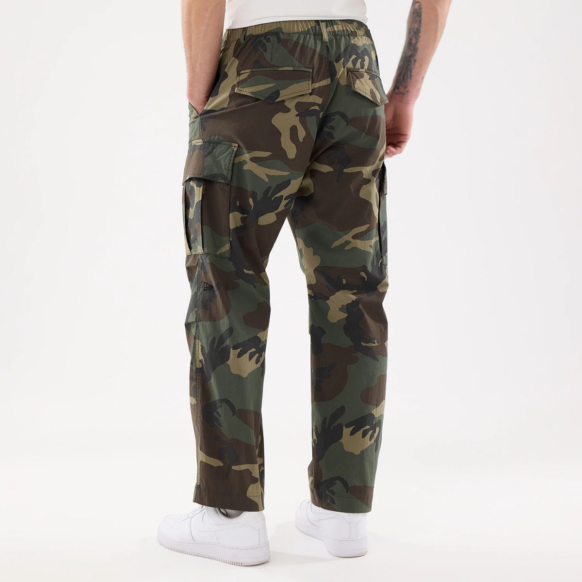 New Era Originators Woodland Camo Cargo Trousers, rear view on a model with hands in pockets, featuring a loose fit design, elastic waistband, and multiple roomy cargo pockets in a woodland camouflage pattern.