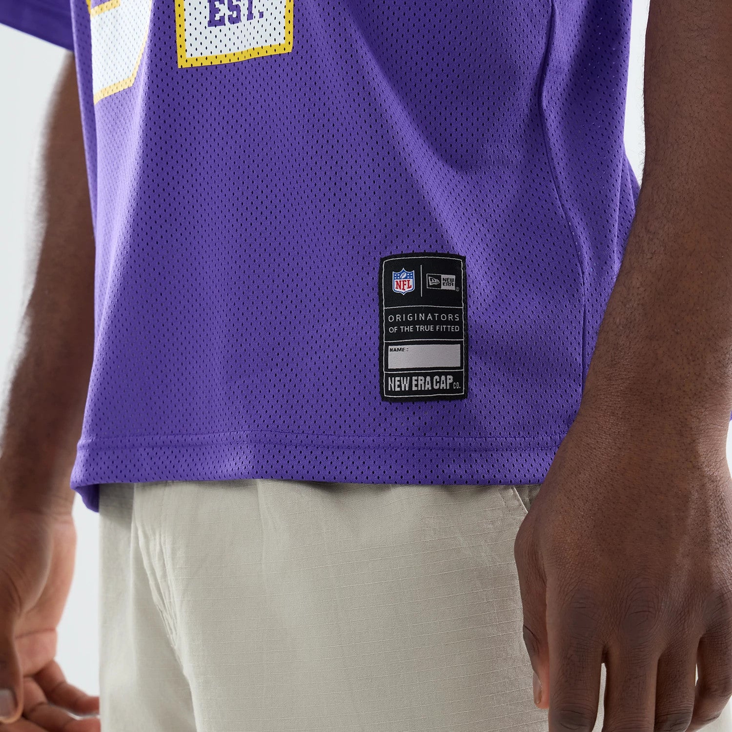 Minnesota Vikings NFL Purple Mesh Jersey, side view on a model showing the lower section with NFL branding and New Era label above the hem, highlighting the perforated fabric texture and loose fit over light trousers.