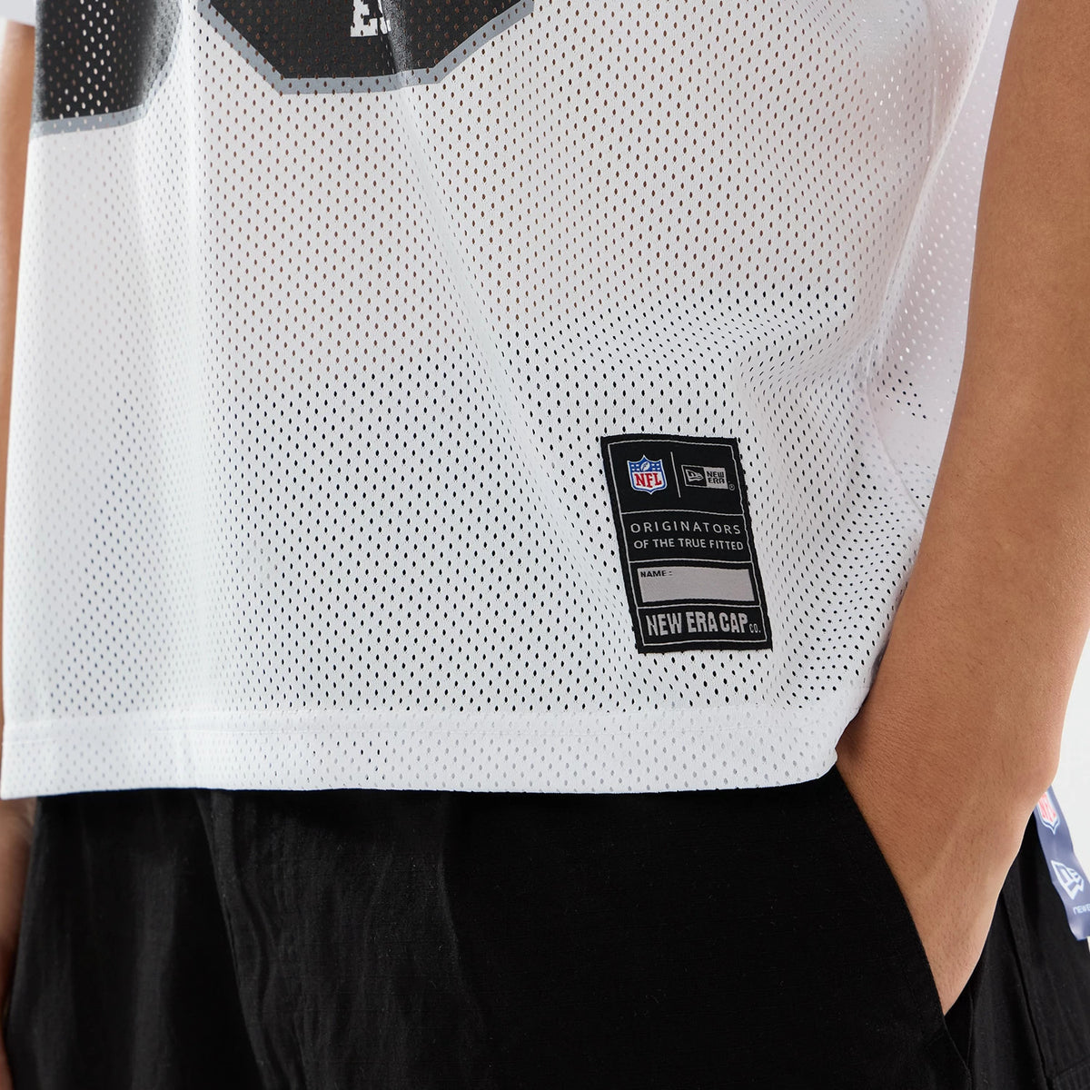 Las Vegas Raiders NFL White Mesh Jersey, close-up showing a black logo patch near the hem, featuring the NFL shield and "ORIGINATORS OF THE TRUE FITTED" text over a breathable perforated fabric.