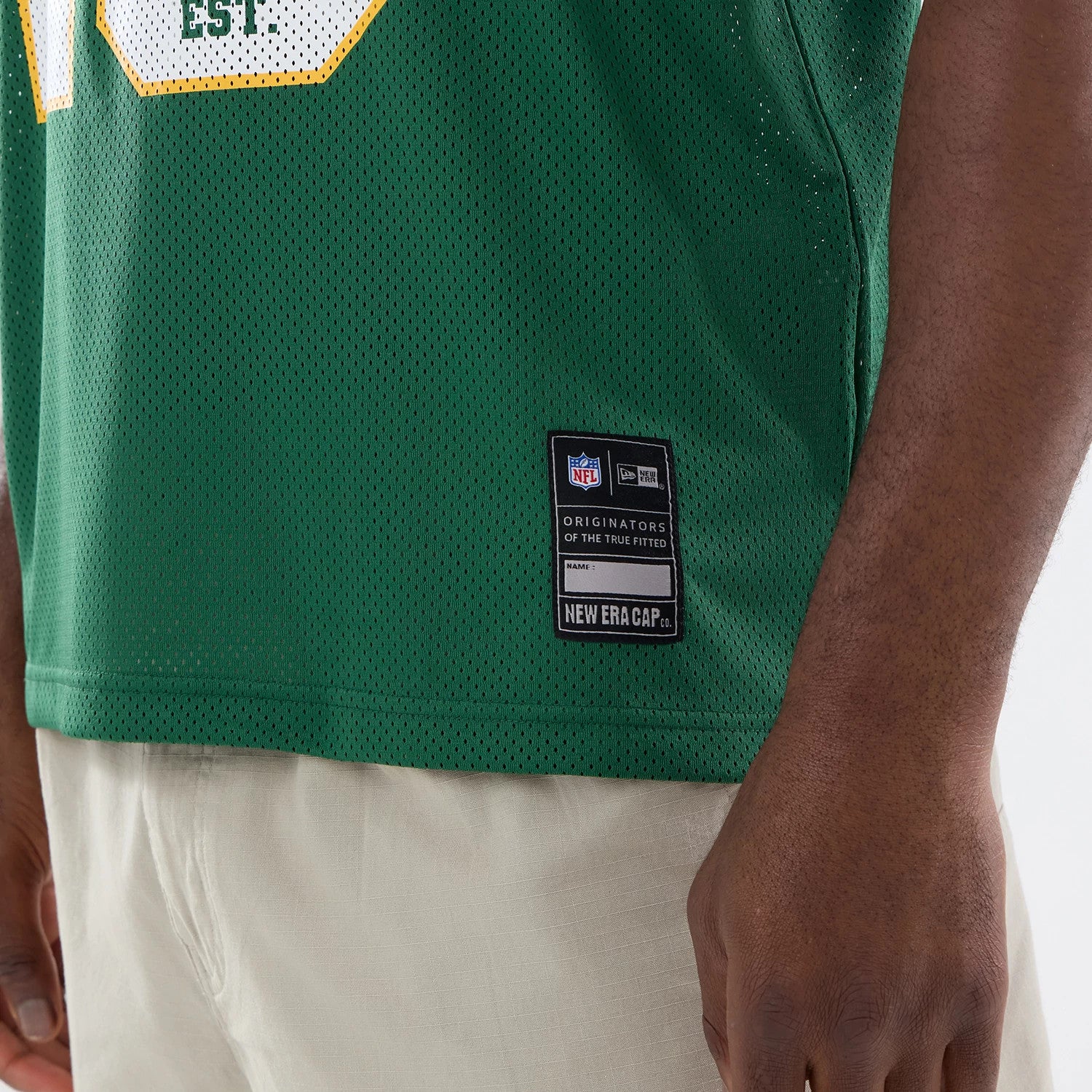 Green Bay Packers NFL Green Mesh Jersey, side view on a model wearing beige trousers showing the stitched black New Era patch with Originators of the True Fitted detail near the hem on the lower side.