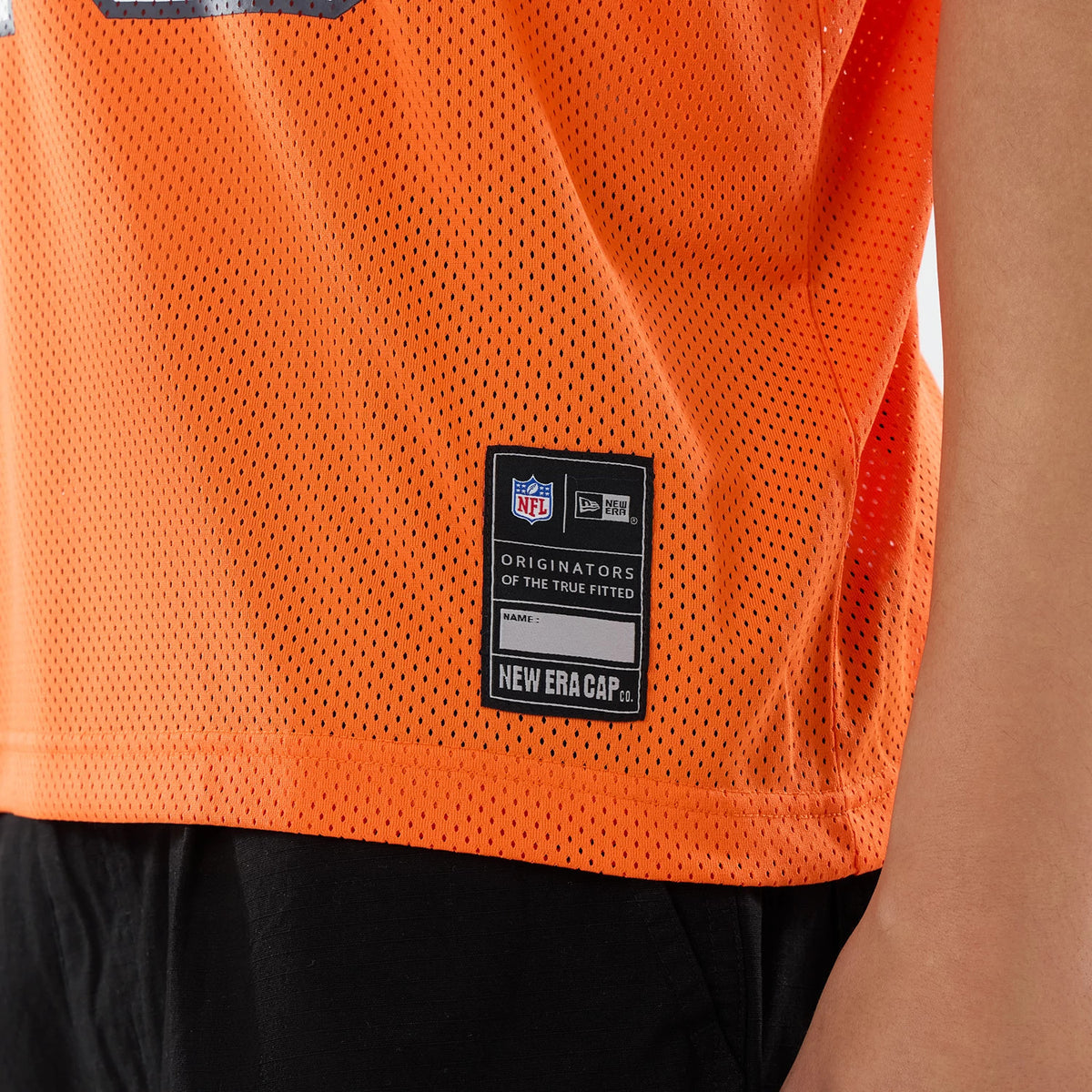 New Era Chicago Bears NFL Orange Mesh Jersey, close-up showing the fabric texture and the sewn-on label at the lower hem with the NFL and New Era logos, worn with black trousers on a model.
