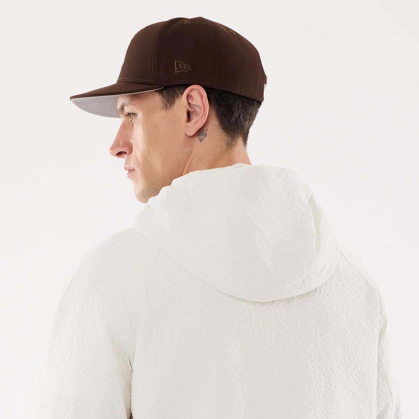 New Era Black Label Open White Jacket, on-model view showing the back of the textured white hooded jacket, worn by a person with a brown cap featuring a debossed New Era logo on the side.