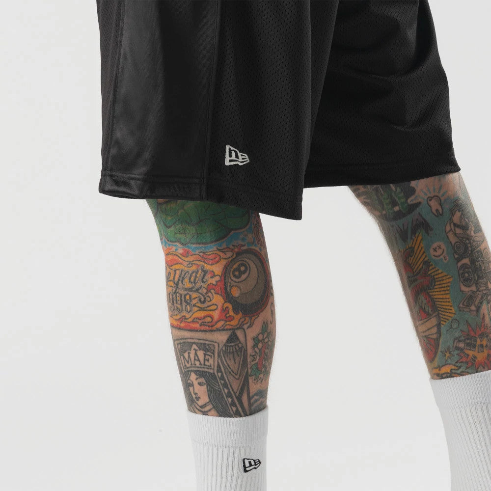 New Era Bermuda Black Oversized Shorts, front view showing the black shorts with a raised New Era logo on the lower right leg, adjacent to colourful tattooed legs and a white sock featuring the New Era logo.