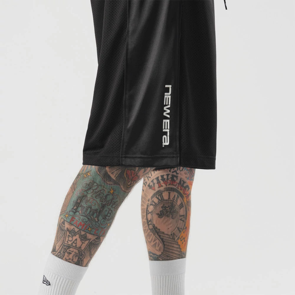 New Era Bermuda Black Oversized Shorts, side view showing the black fabric with a raised white New Era logo running vertically near the edge, worn on tattooed legs above white socks.