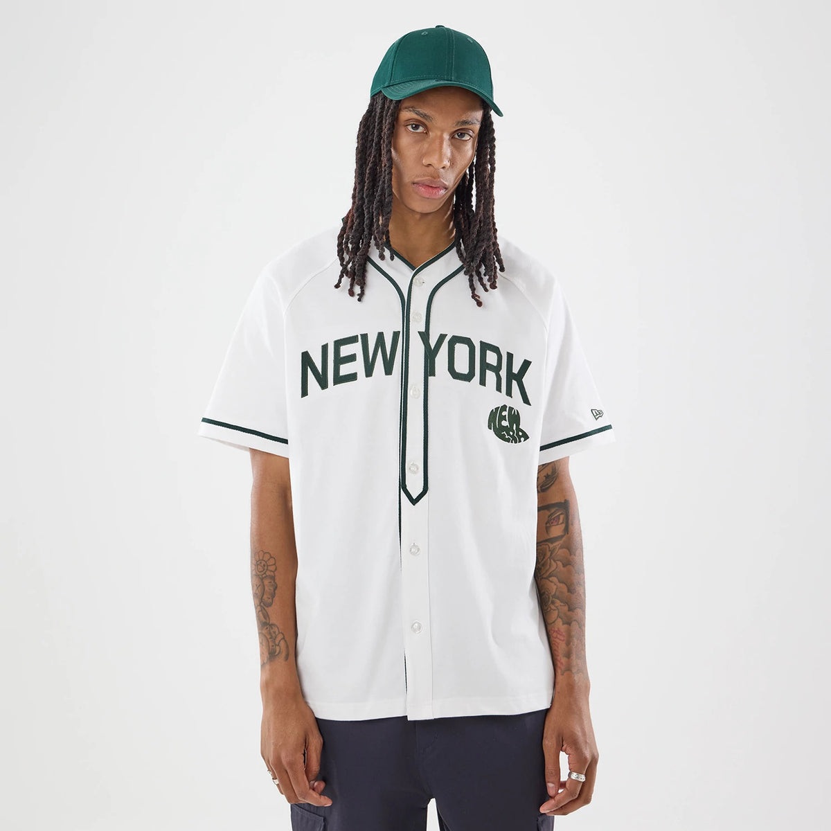 New Era New York White Jersey, front view on a model with a green cap, featuring New York lettering and logo embroidered in dark green on the chest, with black piping on white fabric and a circular graphic on the left side.