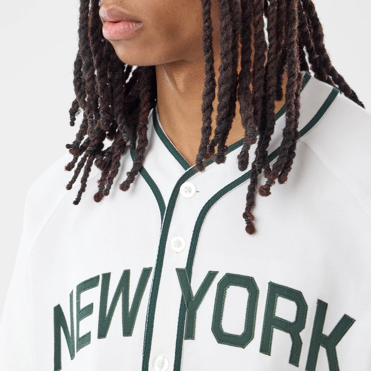 New Era New York White Jersey, front view on a model with extensive dreadlocks, featuring dark green embroidered NEW YORK lettering across the chest, detailed green piping along the button line and white buttons.