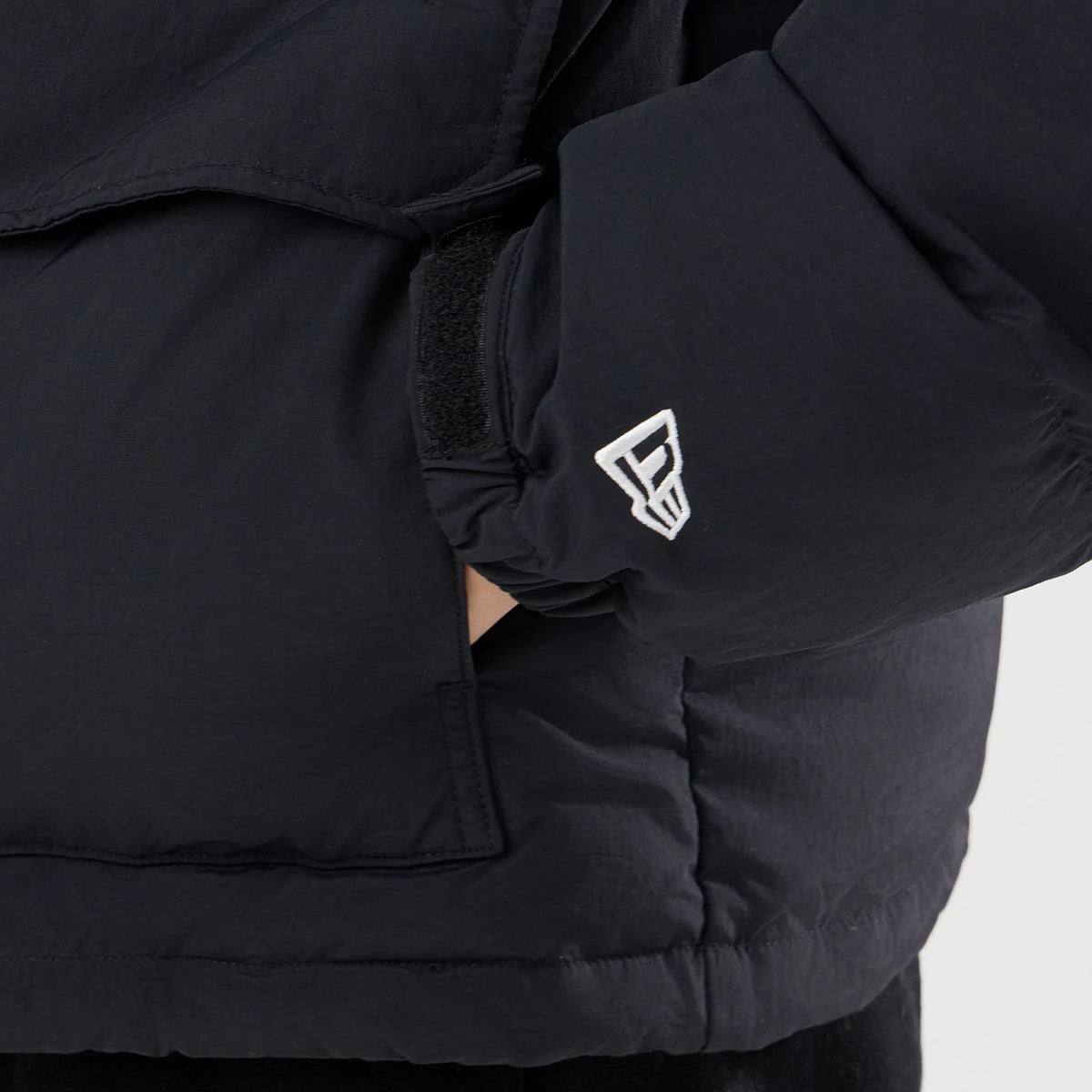 New Era Down Black Jacket, side view showing the black fabric with a white raised New Era logo embroidered on the sleeve near the cuff and a velcro strap for adjustment at the wrist.