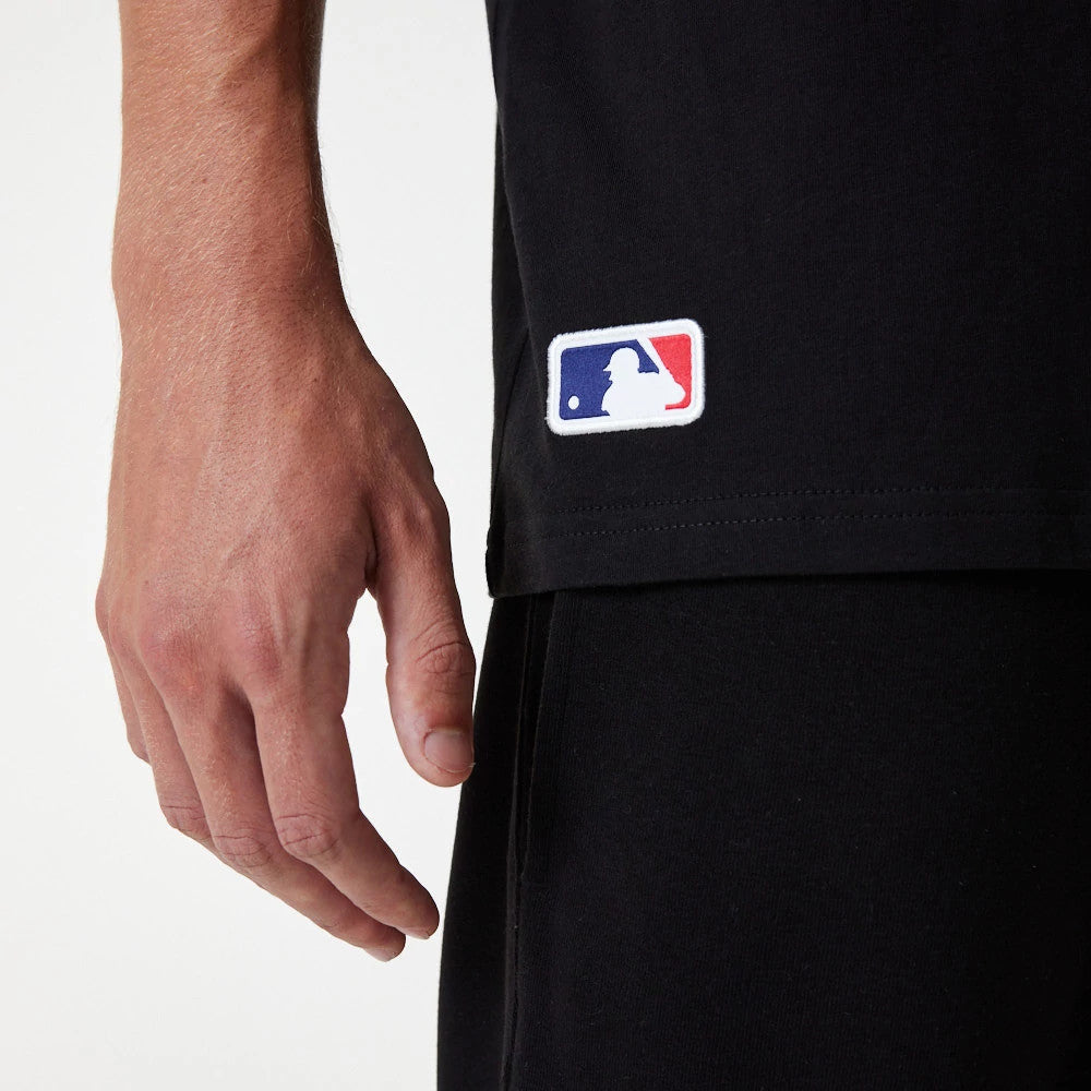 New Era LA Dodgers MLB Essential Black Oversized T-Shirt, side view showing the lower part of the black t-shirt with a multicoloured embroidered MLB logo on the bottom left, set against a black pair of trousers.