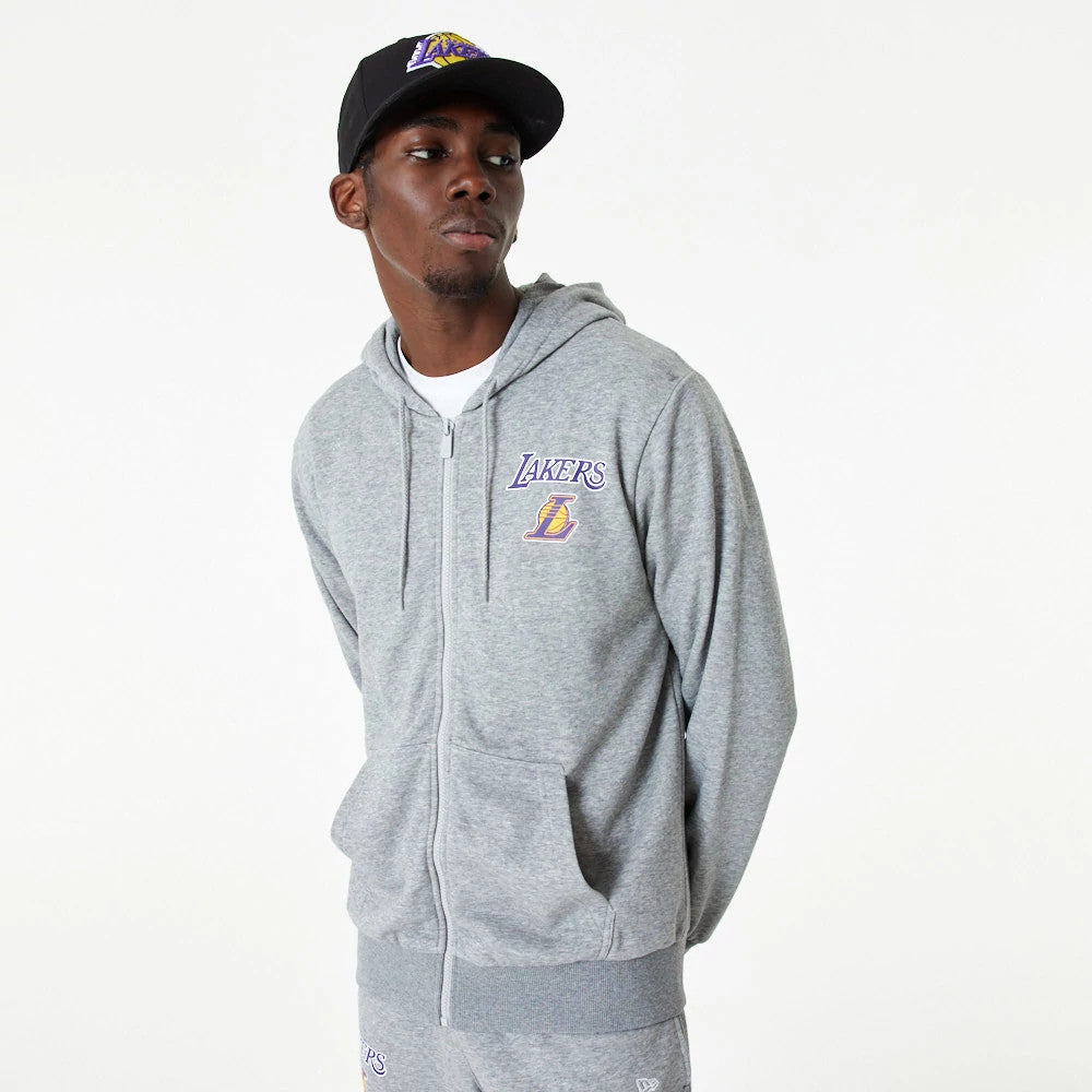 LA Lakers NBA Essential Grey Full Zip Hoodie, on-model view showing the full zip grey hoodie with a raised Lakers logo on the left chest, drawstrings at the hood, and a relaxed fit, worn with matching grey trousers and a black cap with a Lakers logo.