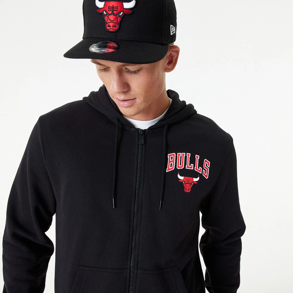Chicago Bulls NBA Essential Black Full Zip Hoodie, on-model view showing the black hoodie with a zip closure down the centre, a red and white Bulls logo embroidered on the left chest, and a raised Bulls logo on a black cap.