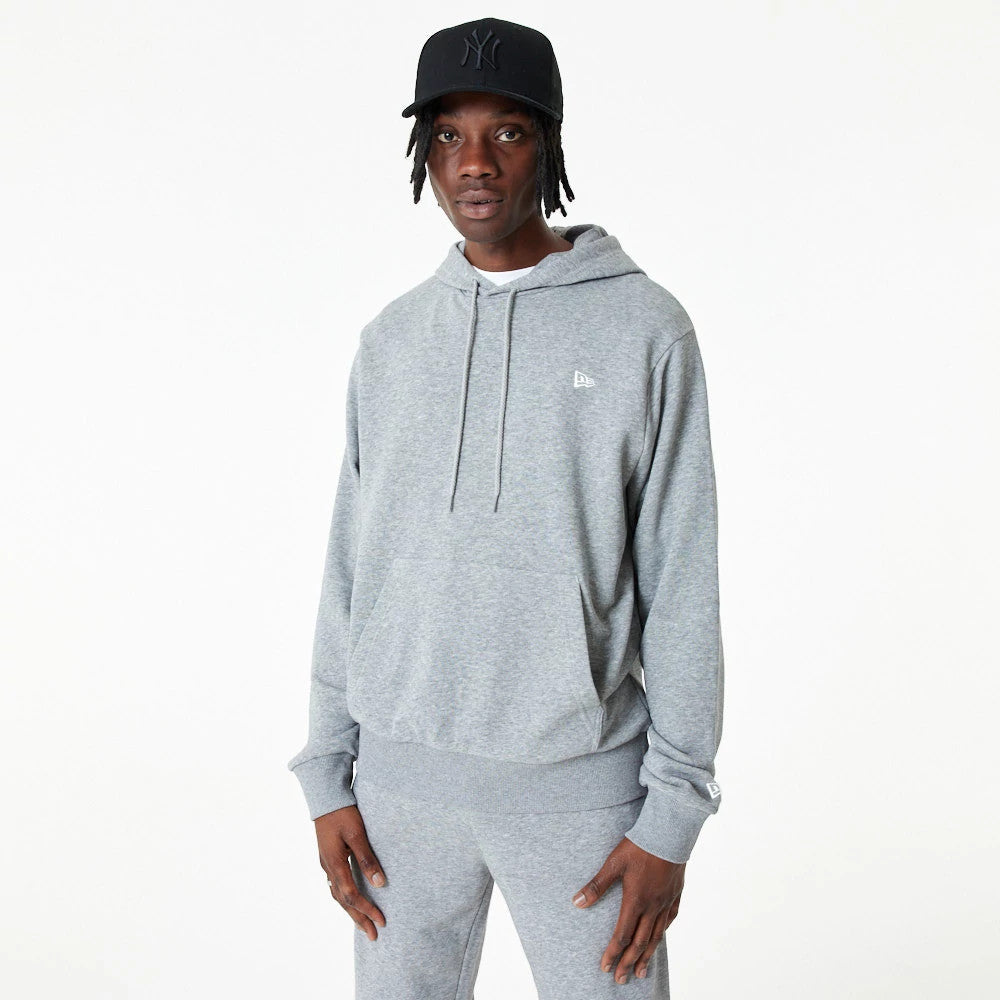 New Era Essential Grey Pullover Hoodie, on-model view showing a light grey hoodie with a raised New Era logo on the chest, adjustable drawstrings, and matching grey trousers, paired with a black cap featuring a logo on the front.