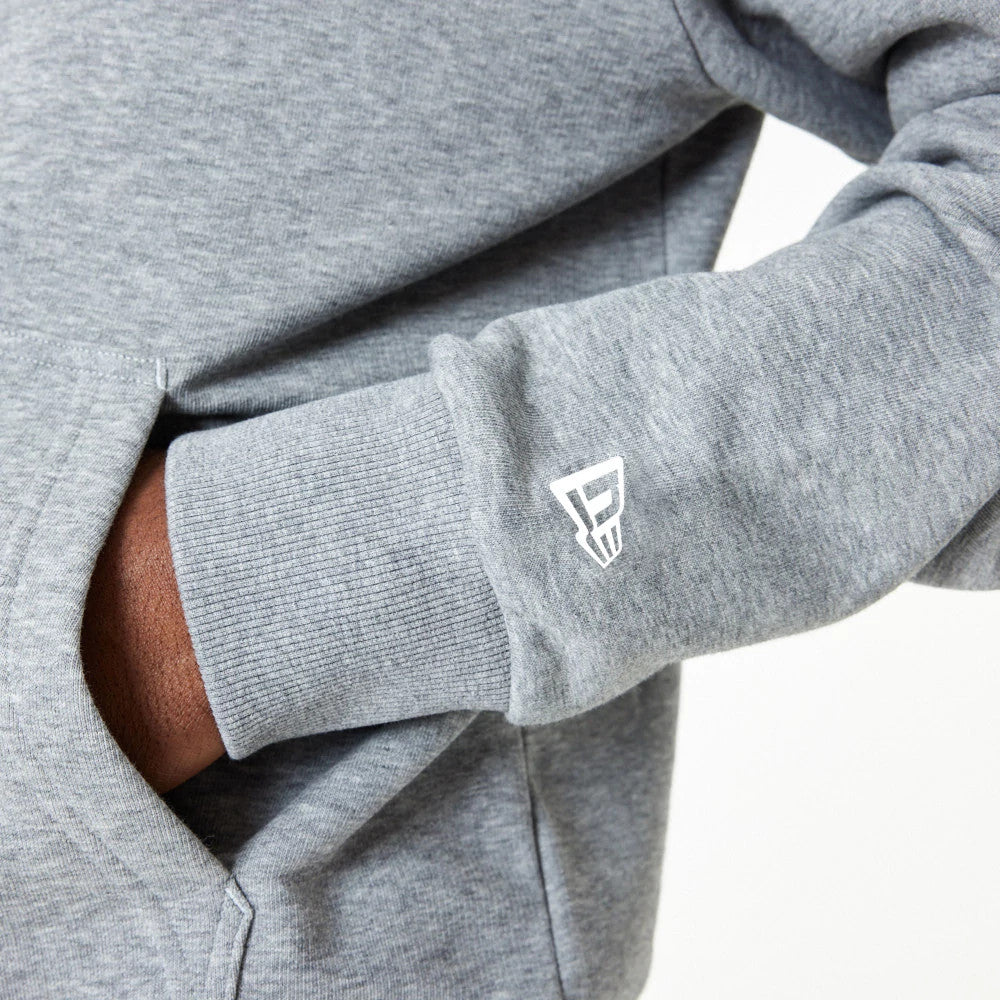 New Era Essential Grey Pullover Hoodie, side view showing a close-up of the grey fabric sleeve with ribbed cuff, a white embossed New Era logo on the wrist, and the arm partially covering the front pouch pocket.