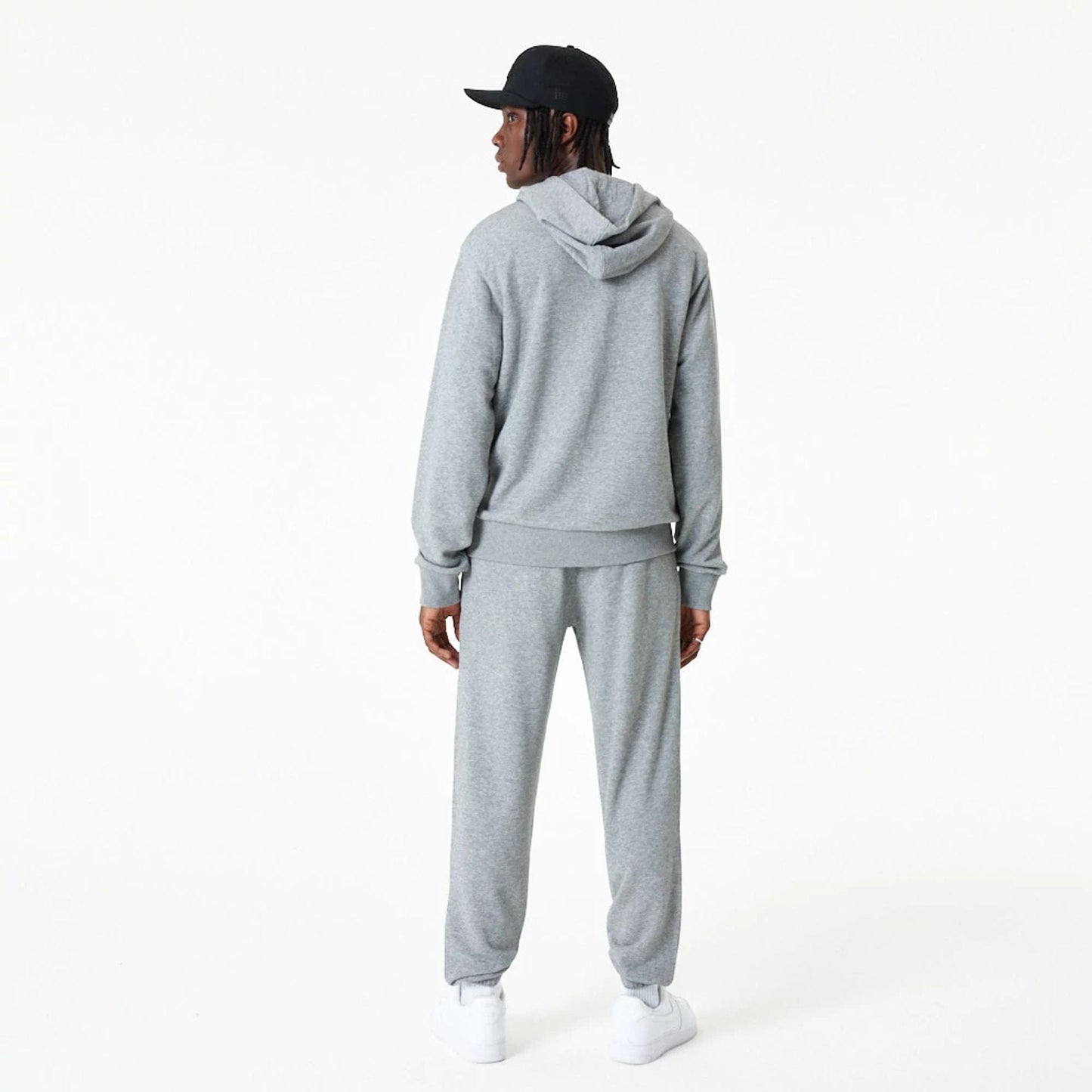 New Era Essential Grey Pullover Hoodie, rear view showing the relaxed fit with a drawstring hood, ribbed cuffs, and hem, worn by a person also wearing matching grey pants and a black cap with a debossed logo.